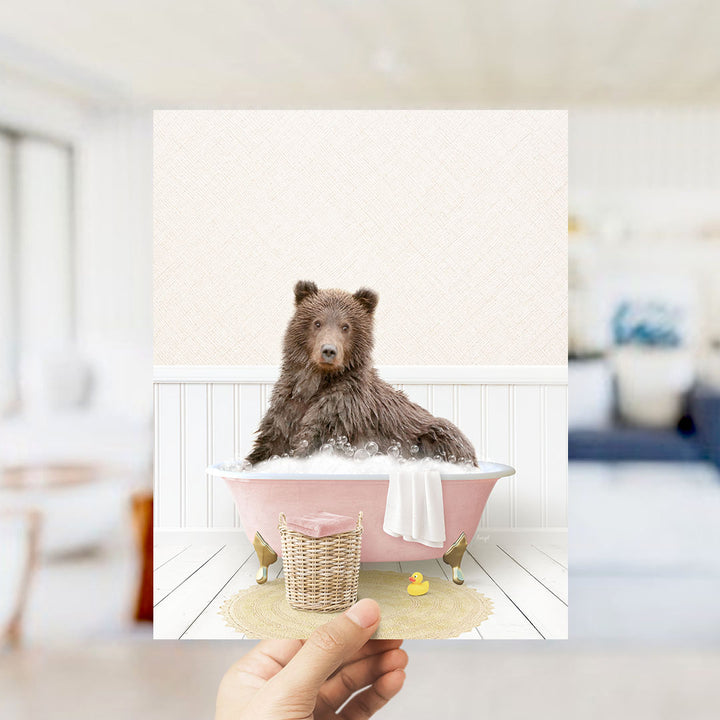 A brown bear sitting in a pink bathtub filled with bubbles, surrounded by a yellow rubber duck and a white towel.