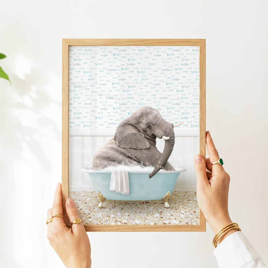 A person's hands are holding a picture frame containing an image of a gray elephant sitting in a blue bathtub filled with water and bubbles.