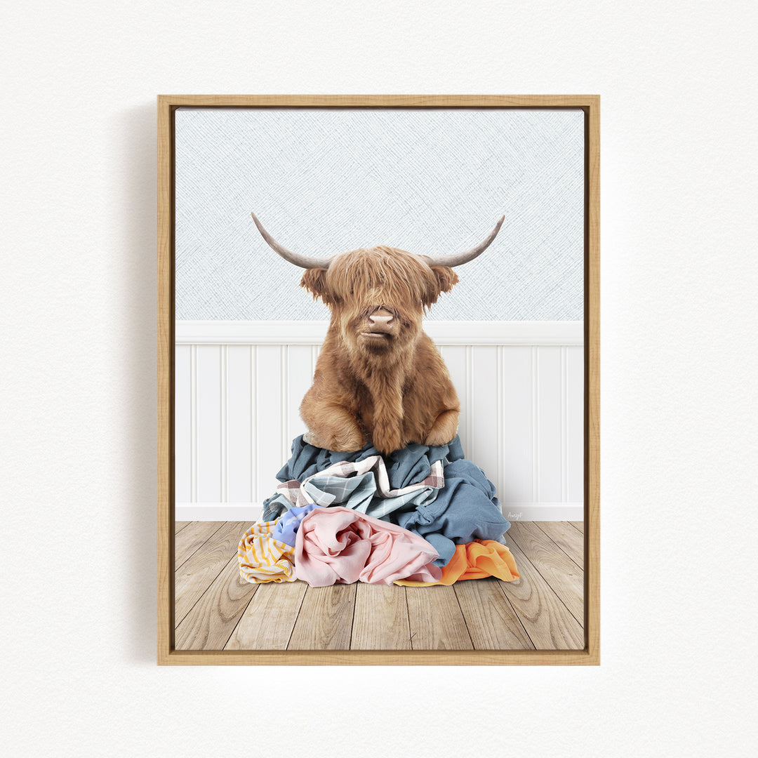 A Highland cow with long horns is sitting atop a pile of clothes on a wooden floor.