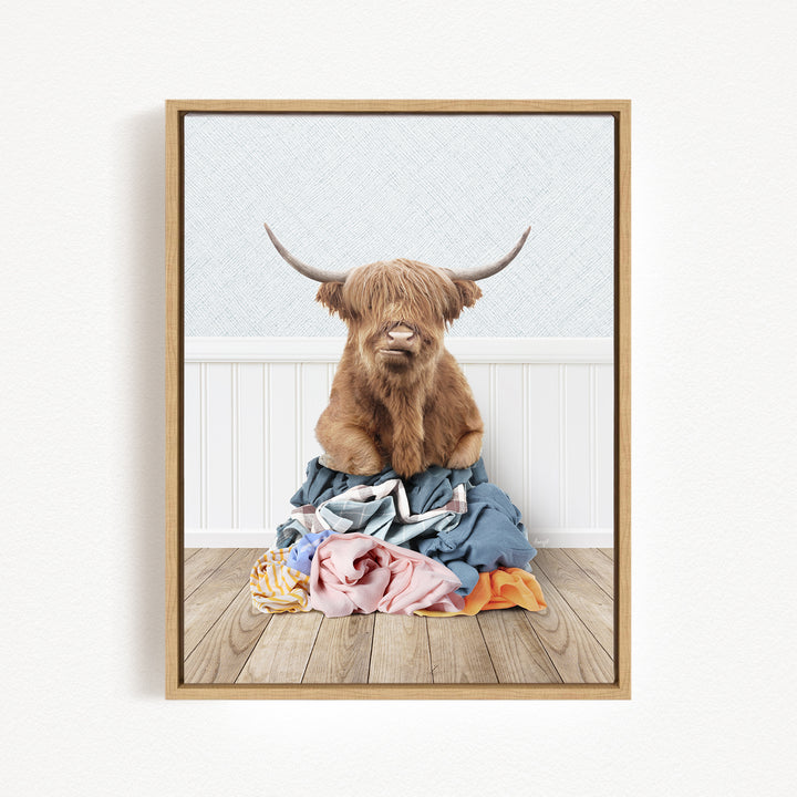 A Highland cow with long horns is sitting atop a pile of clothes on a wooden floor.