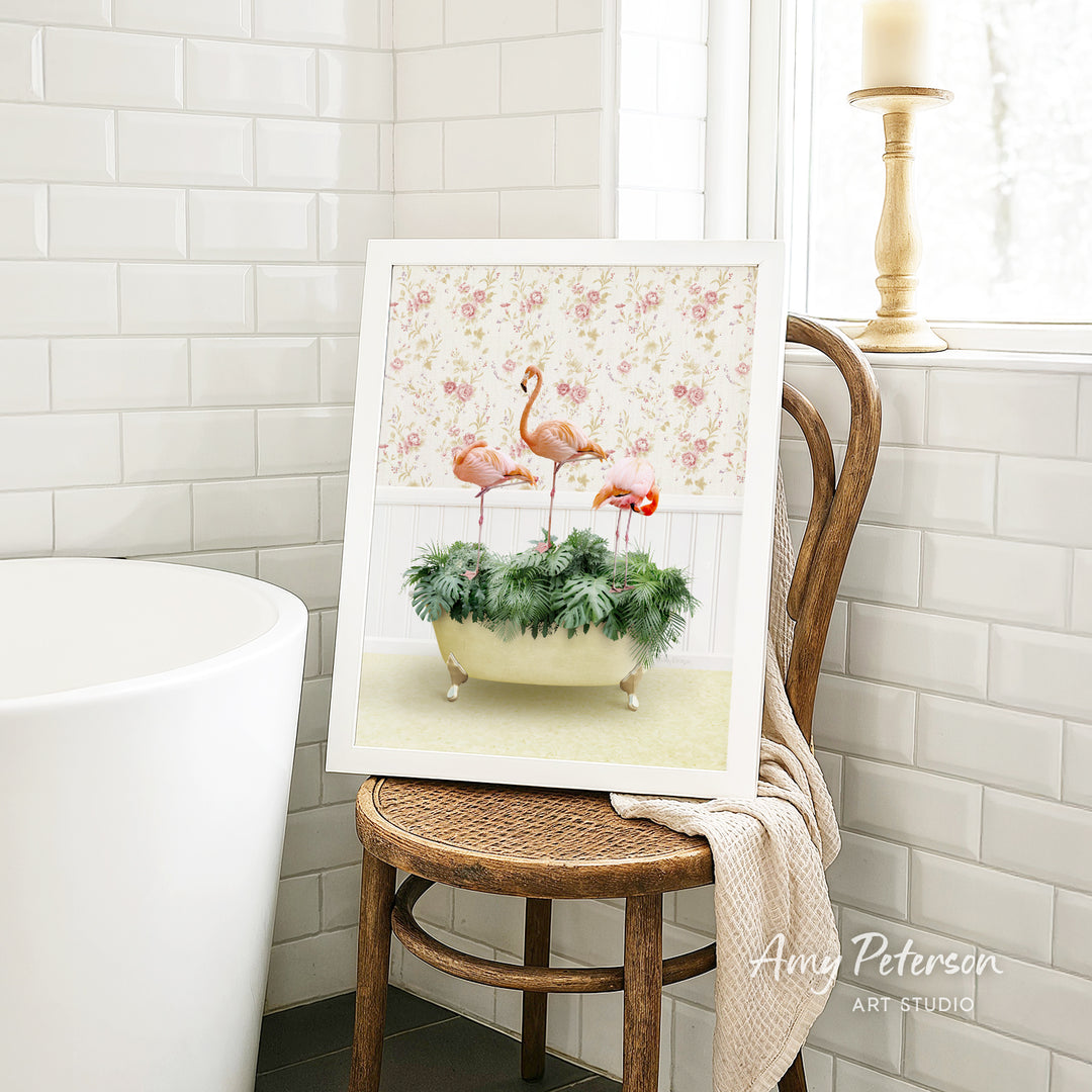 A framed image of three flamingos standing in a bathtub filled with green plants is displayed on a wooden chair in a bathroom setting.