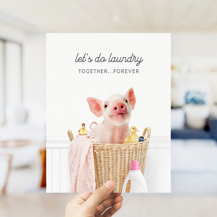 A hand holding a greeting card featuring a pig sitting in a wicker basket filled with various household items, including a bottle of detergent and a yellow duck.