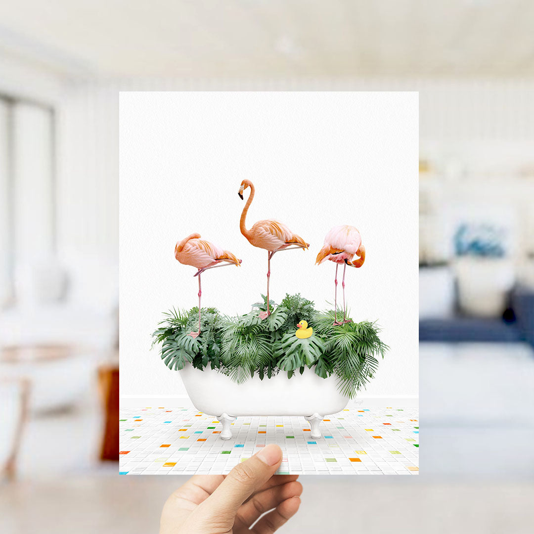 A hand holds up a framed image depicting three pink flamingos standing in a bathtub filled with green plants and a yellow rubber duck.