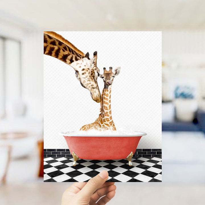 A hand holds up a framed image depicting two giraffes in a red bathtub, with one giraffe appearing to be bathing the other.
