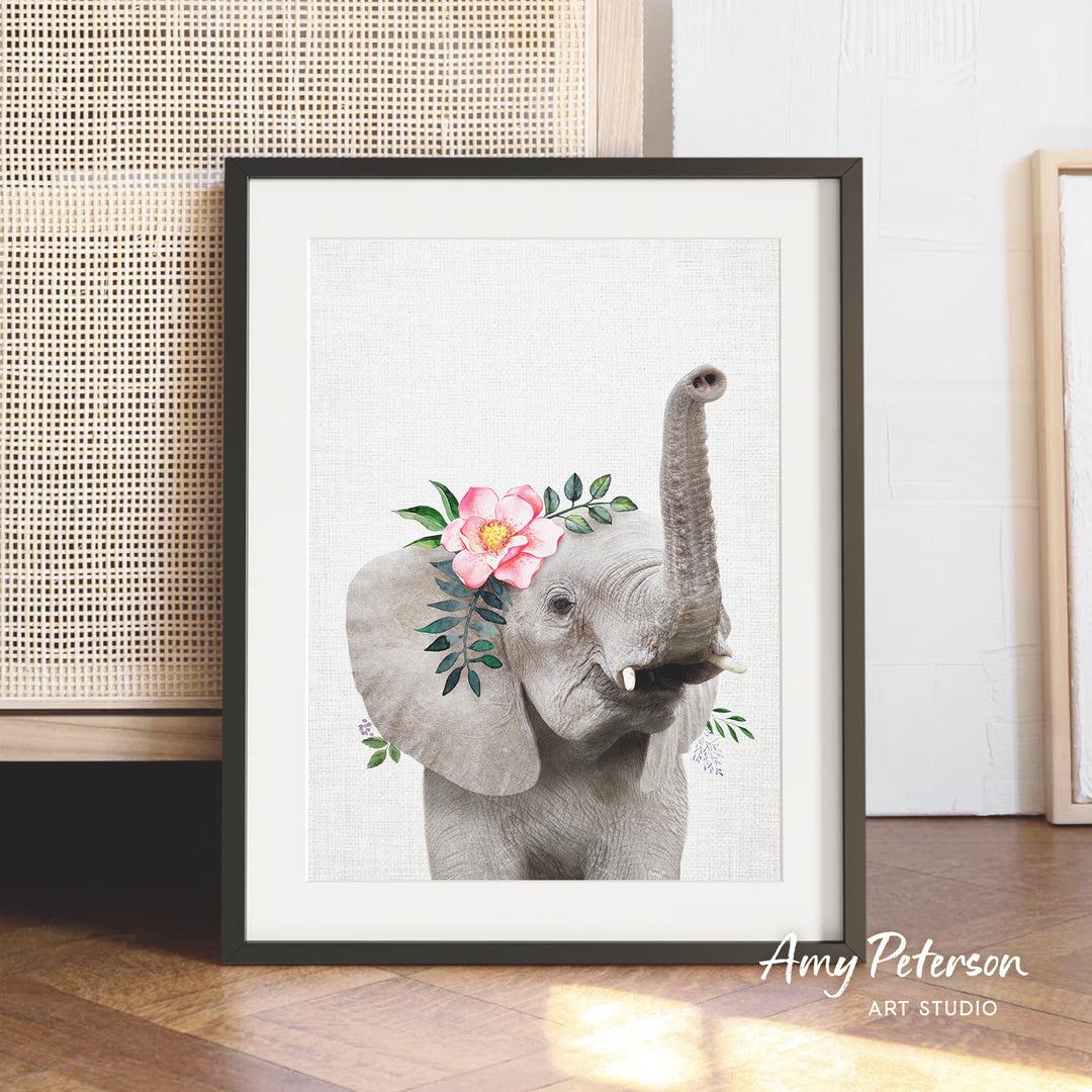 A framed print of an elephant with a pink flower on its head is displayed on a wooden floor.