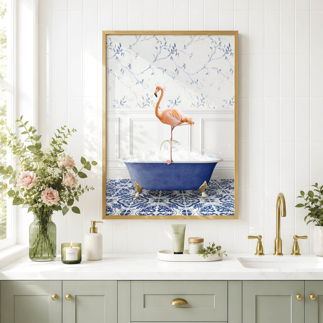 a framed painting of a pink flamingo standing in a blue bathtub, surrounded by a floral pattern on the wall. The painting is hung above a white countertop with various bathroom accessories, including a vase of pink flowers, a soap dispenser, and a candle.
