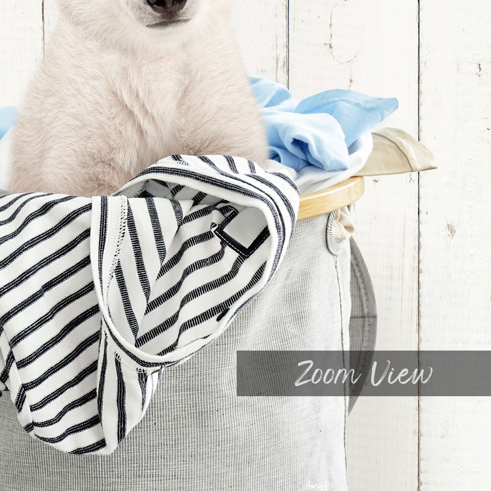 A white dog is sitting in a basket filled with clothes, with a wooden wall in the background.