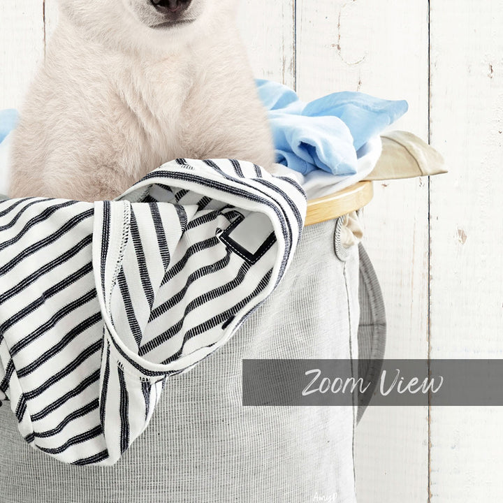 A white dog is sitting in a basket filled with clothes, with a wooden wall in the background.