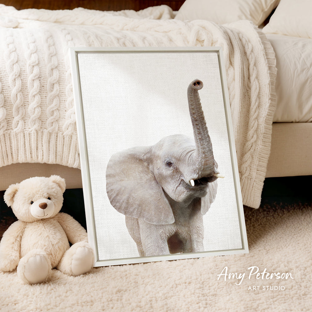 A framed photograph of a baby elephant is displayed next to a teddy bear on a bed, with a white blanket and a wooden headboard in the background.