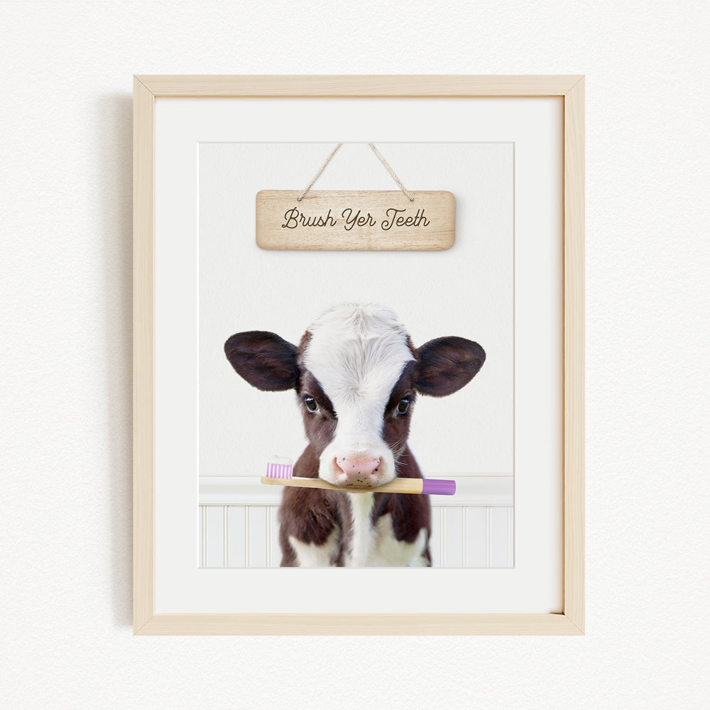 A framed image of a cow holding a purple toothbrush in its mouth, with a wooden sign above it that says "Brush Your Teeth".