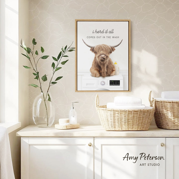 a framed poster of a Highland cow with horns, hanging on a wall above a white cabinet with baskets and a vase of leaves.