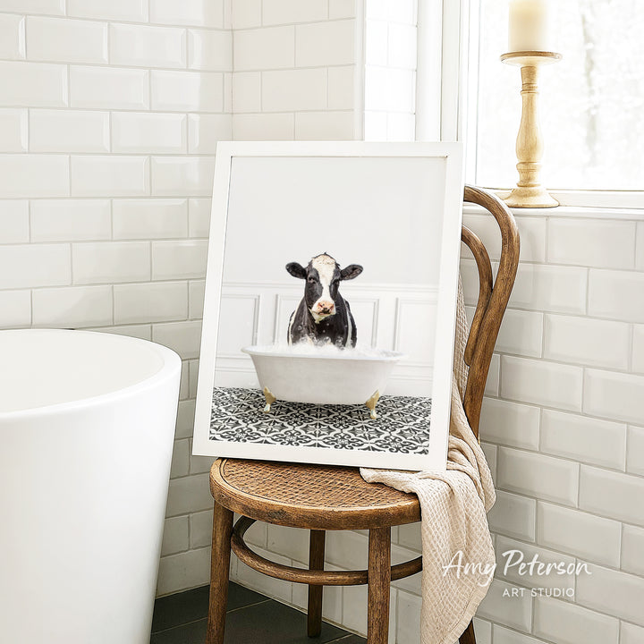 A framed image of a cow sitting in a bathtub, with a towel draped over the back of a wooden chair in a bathroom setting.