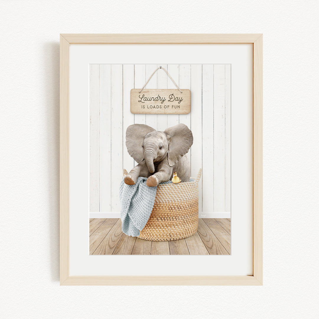 A framed image of a baby elephant sitting in a woven basket, with a sign above that says "Laundry Day is loads of fun".