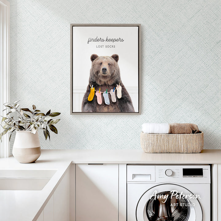 a framed poster of a bear hanging clothes on a clothesline, with the text "Finders keepers" and "Lost socks" written above it. The poster is displayed on a wall above a white countertop, which has a potted plant and a basket of towels on it.