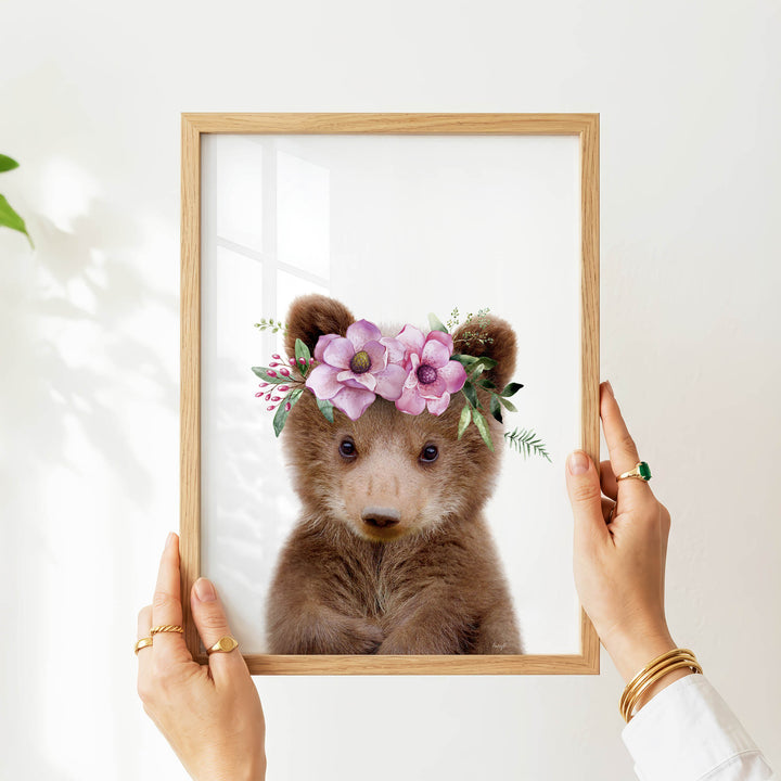 A person's hands are holding a wooden frame with a picture of a brown bear wearing a floral crown.