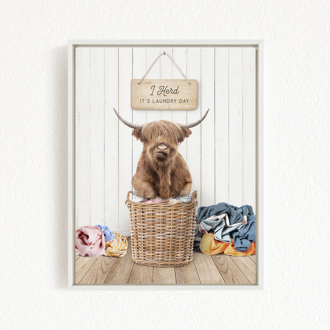 A Highland cow sits in a wicker basket filled with laundry, with a sign above that says "It's laundry day".