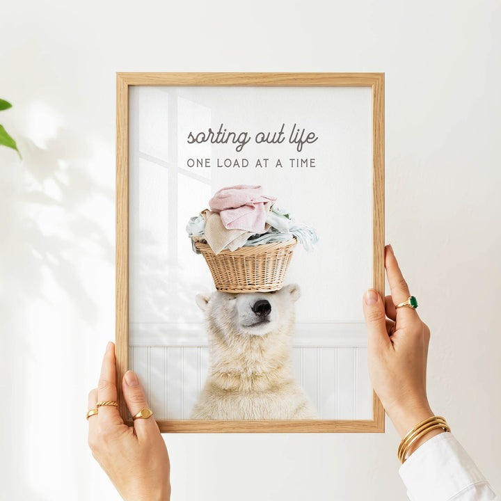 A person is holding up a framed image of a polar bear wearing a basket on its head, with the text "sorting out life one load at a time" written above the bear.