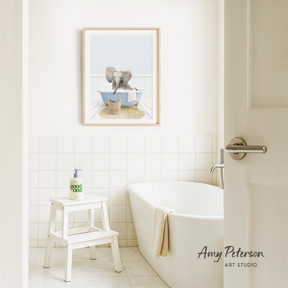 A framed image of an elephant sitting in a bathtub is displayed on the wall above a white bathtub in a bathroom.