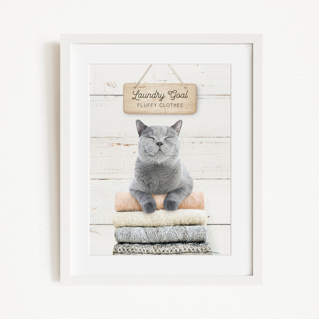 A gray cat sitting on a stack of folded clothes, with a sign above that says "Laundry Coal Fluffy Clothes".
