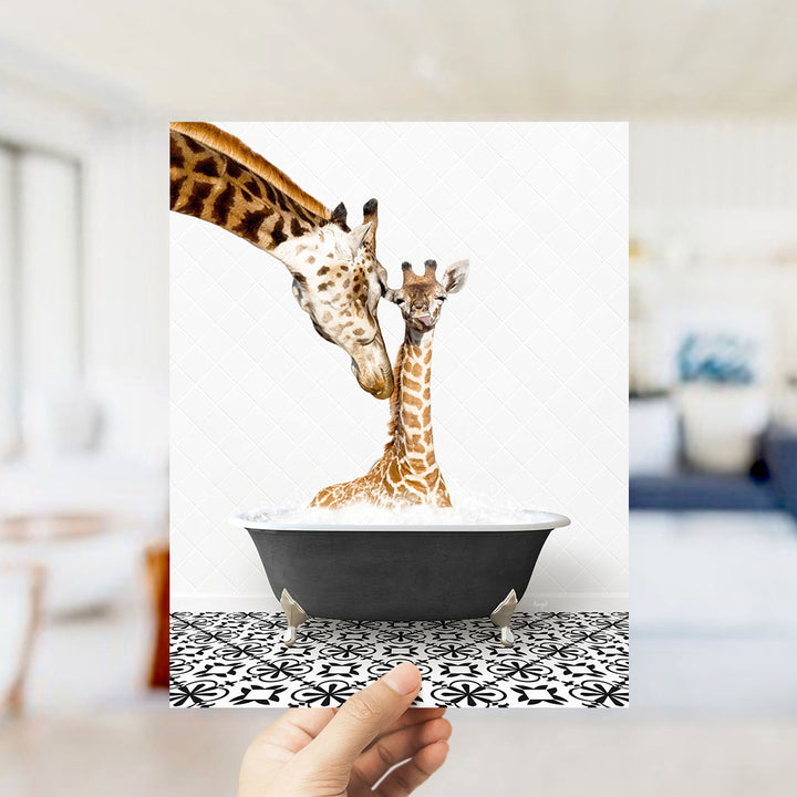 A hand holds up a framed photograph of two giraffes in a bathtub, with one giraffe appearing to be a baby and the other an adult.