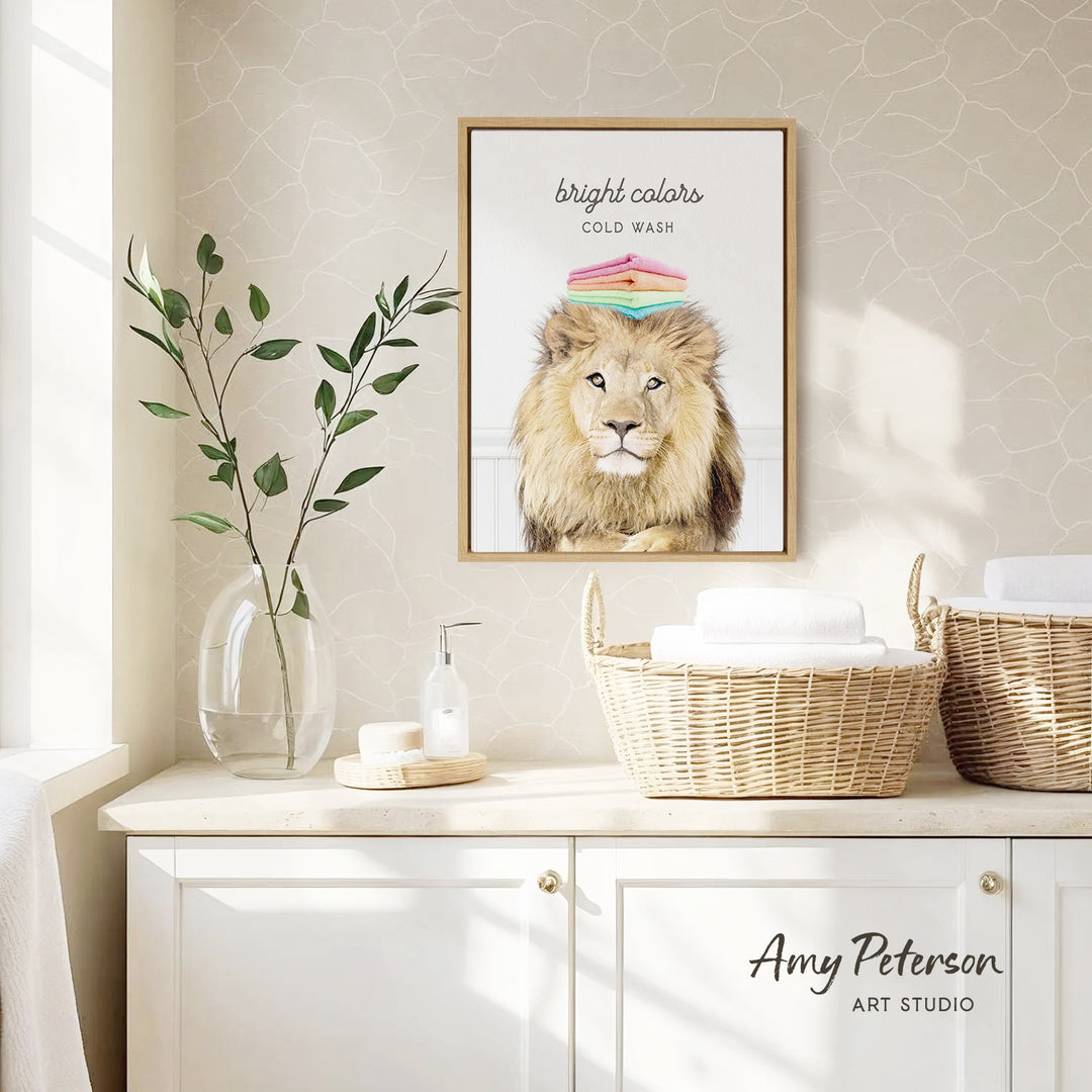a framed poster of a lion with a colorful hat on its head, hanging on a wall above a white cabinet. The cabinet has two woven baskets on top, and there is a vase with a plant on the counter. The wall has a patterned wallpaper, and the overall scene has a clean and minimalist aesthetic.