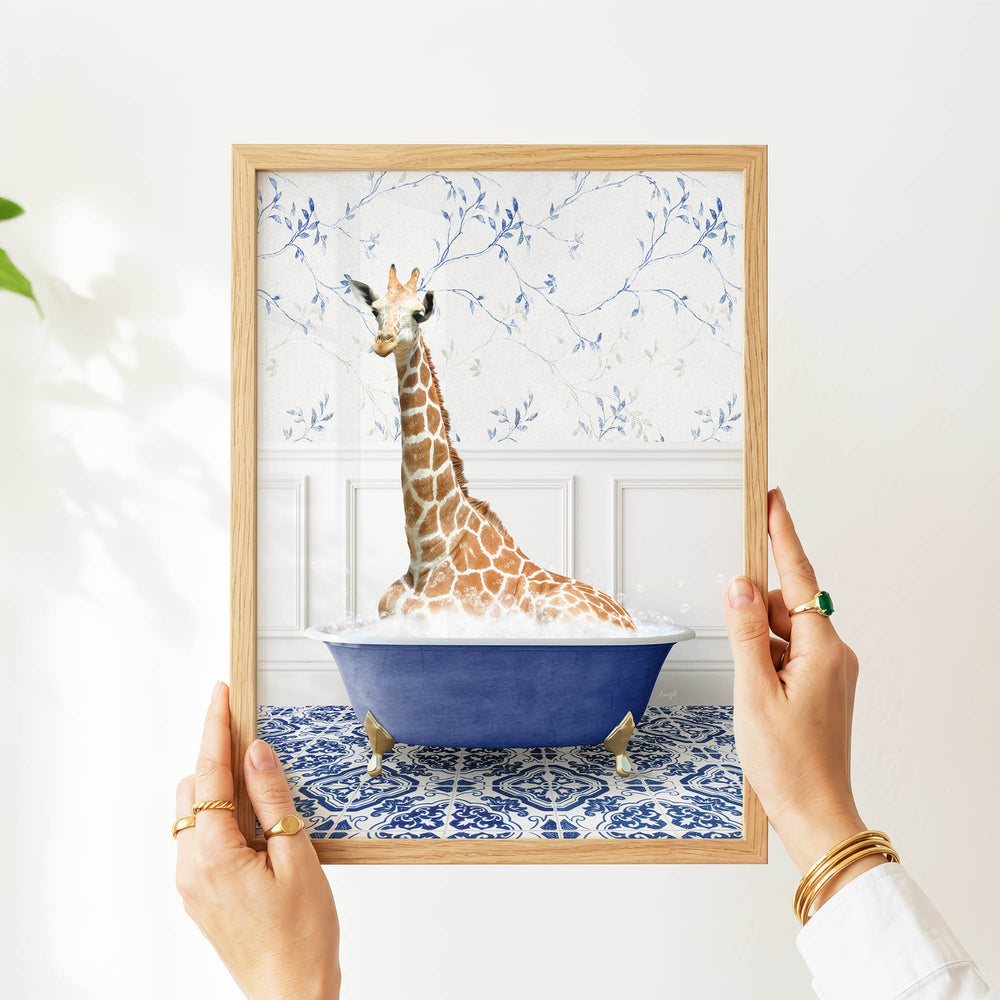 A person's hands are holding a framed picture of a giraffe sitting in a blue bathtub, with a patterned wall behind it.