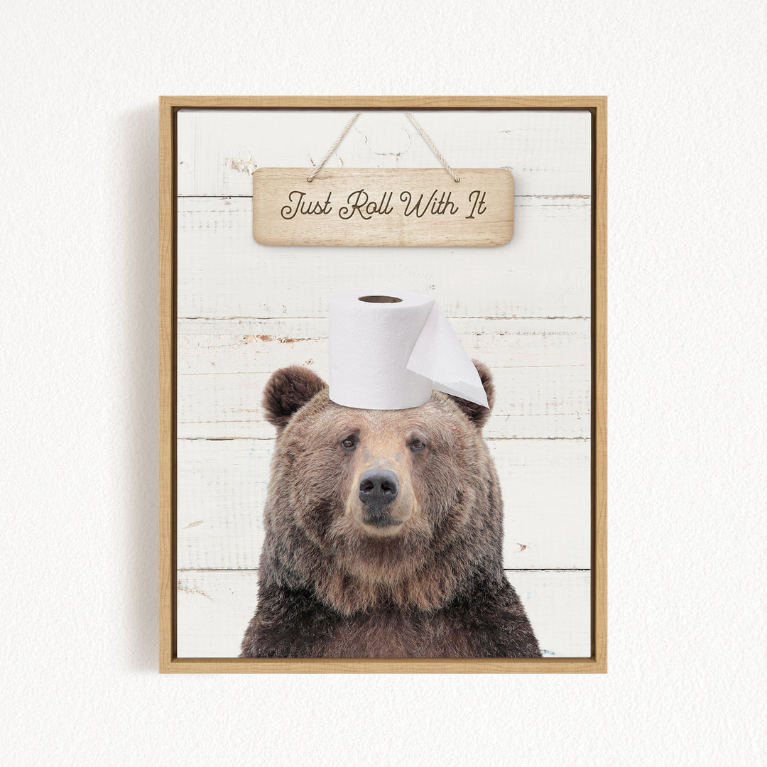 A framed image of a brown bear wearing a roll of toilet paper on its head, with a wooden sign above that says "Just Roll With It".
