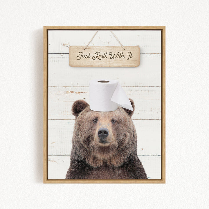 A framed image of a brown bear wearing a roll of toilet paper on its head, with a wooden sign above that says "Just Roll With It".
