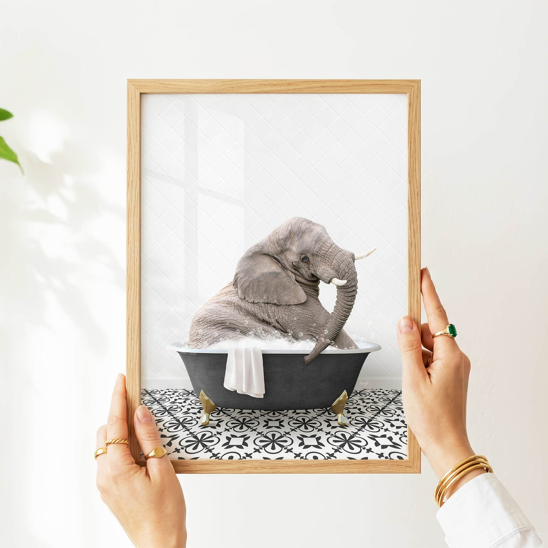 A person's hands are holding a picture frame containing an image of a gray elephant sitting in a bathtub filled with water and bubbles.