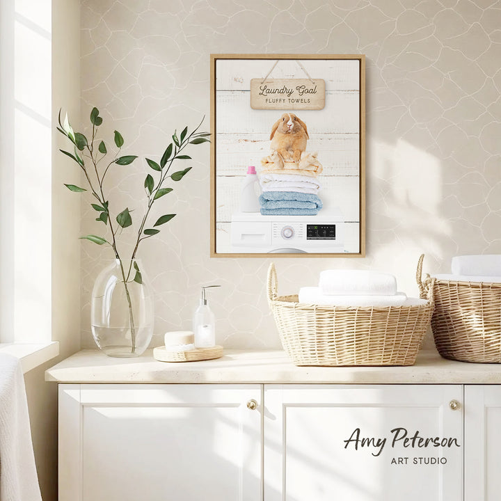 a framed picture of a fluffy towel hanging on a wall, with a wicker basket below it. The basket is filled with folded towels. There is also a vase with a plant and a bottle of soap on a countertop.