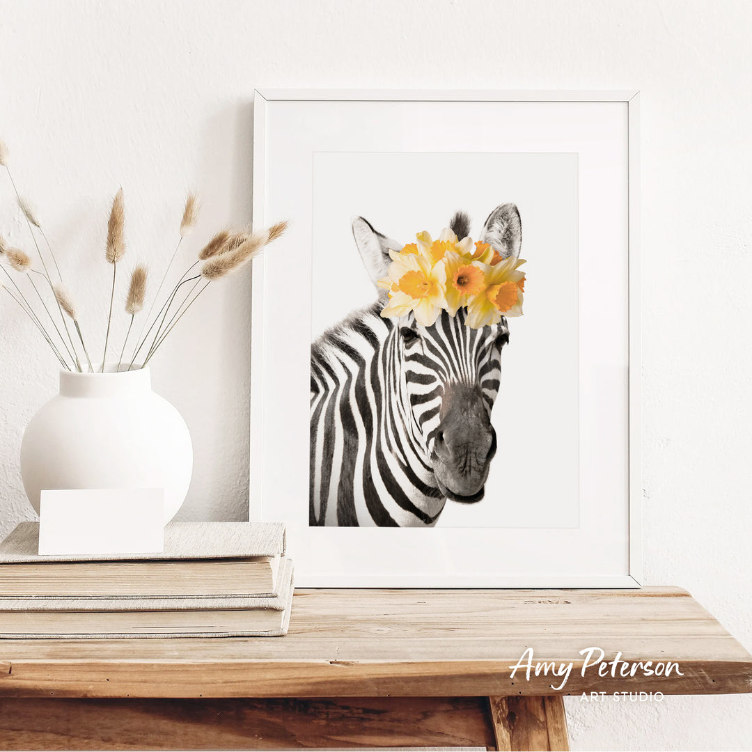 A framed photograph of a zebra with yellow flowers on its head is displayed on a wooden shelf, accompanied by a white vase with dried flowers and a stack of books.