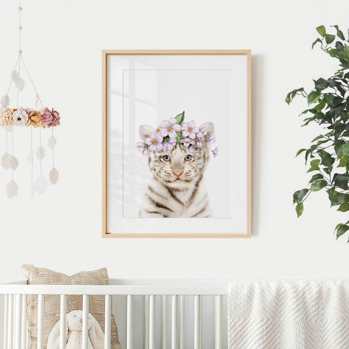 A framed photograph of a tiger cub wearing a flower crown is displayed on a white wall above a white crib.