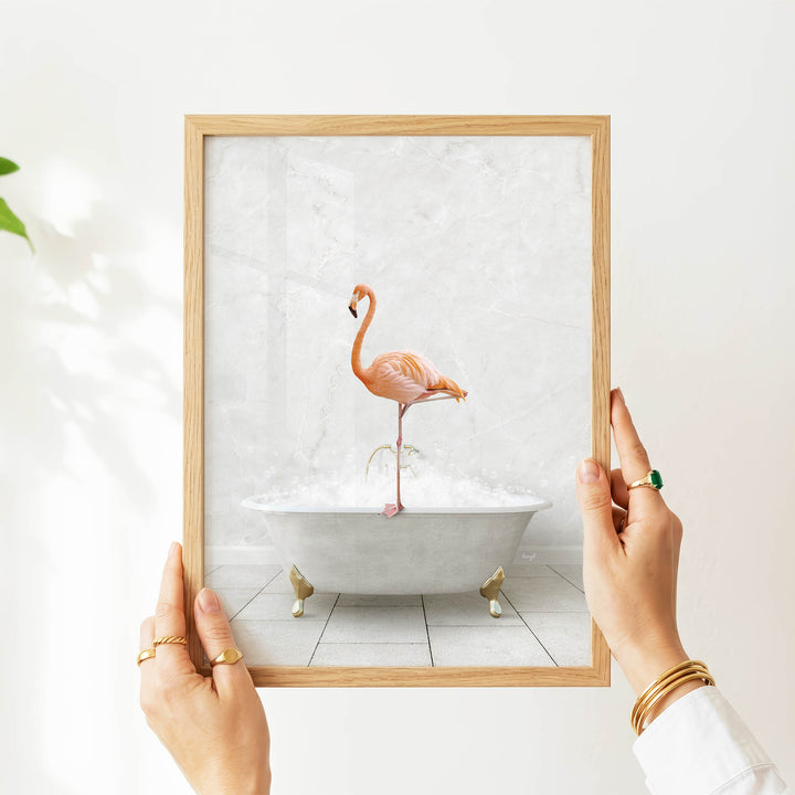A person's hands are holding a framed picture of a flamingo standing in a bathtub filled with bubbles.