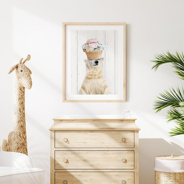 a wooden dresser with a framed picture of a polar bear wearing a basket on its head, placed above it. Next to the dresser is a stuffed giraffe toy. The room also has a potted plant and a white wall.