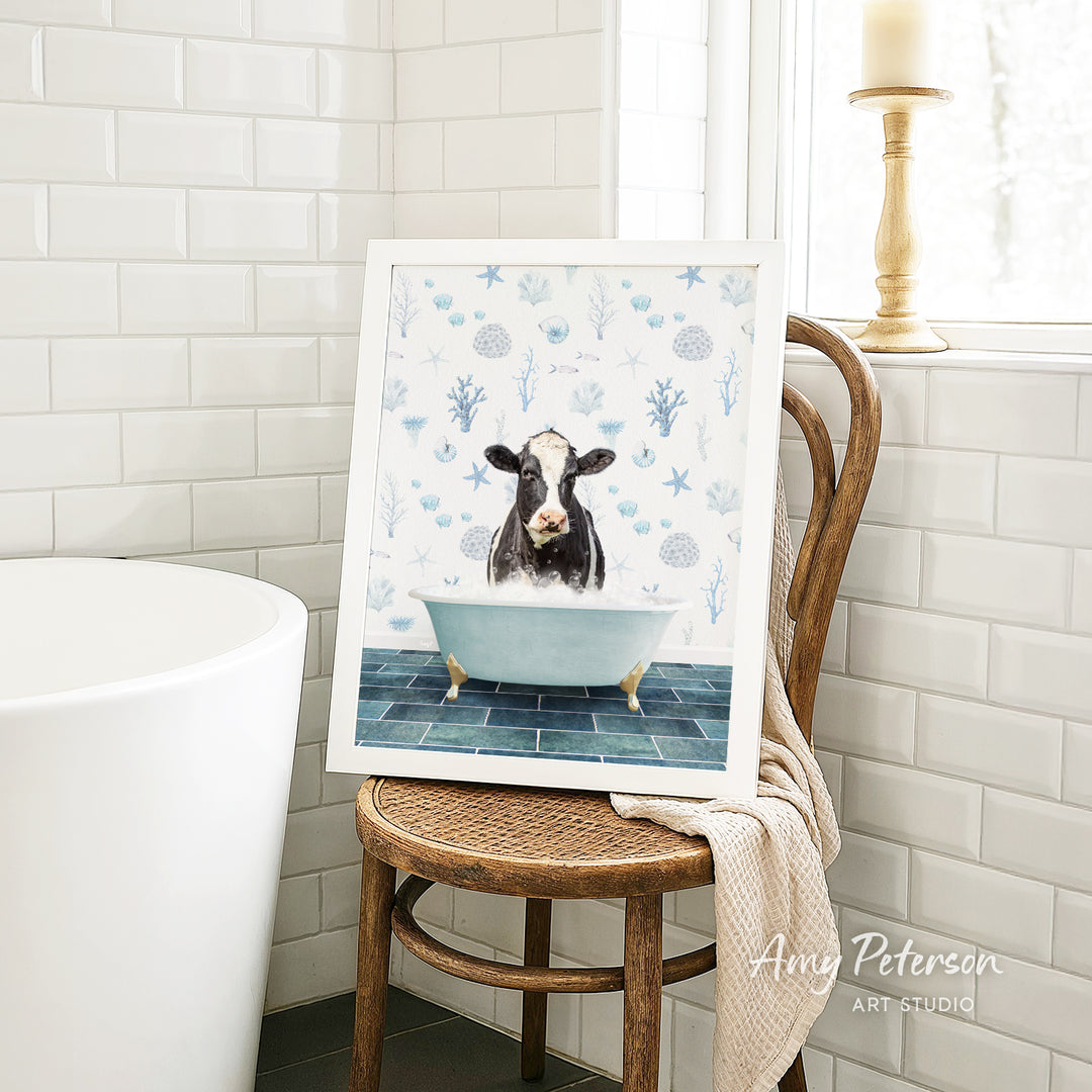 A framed image of a cow sitting in a bathtub, surrounded by blue and white floral patterns, is displayed on a wooden chair in a bathroom setting.