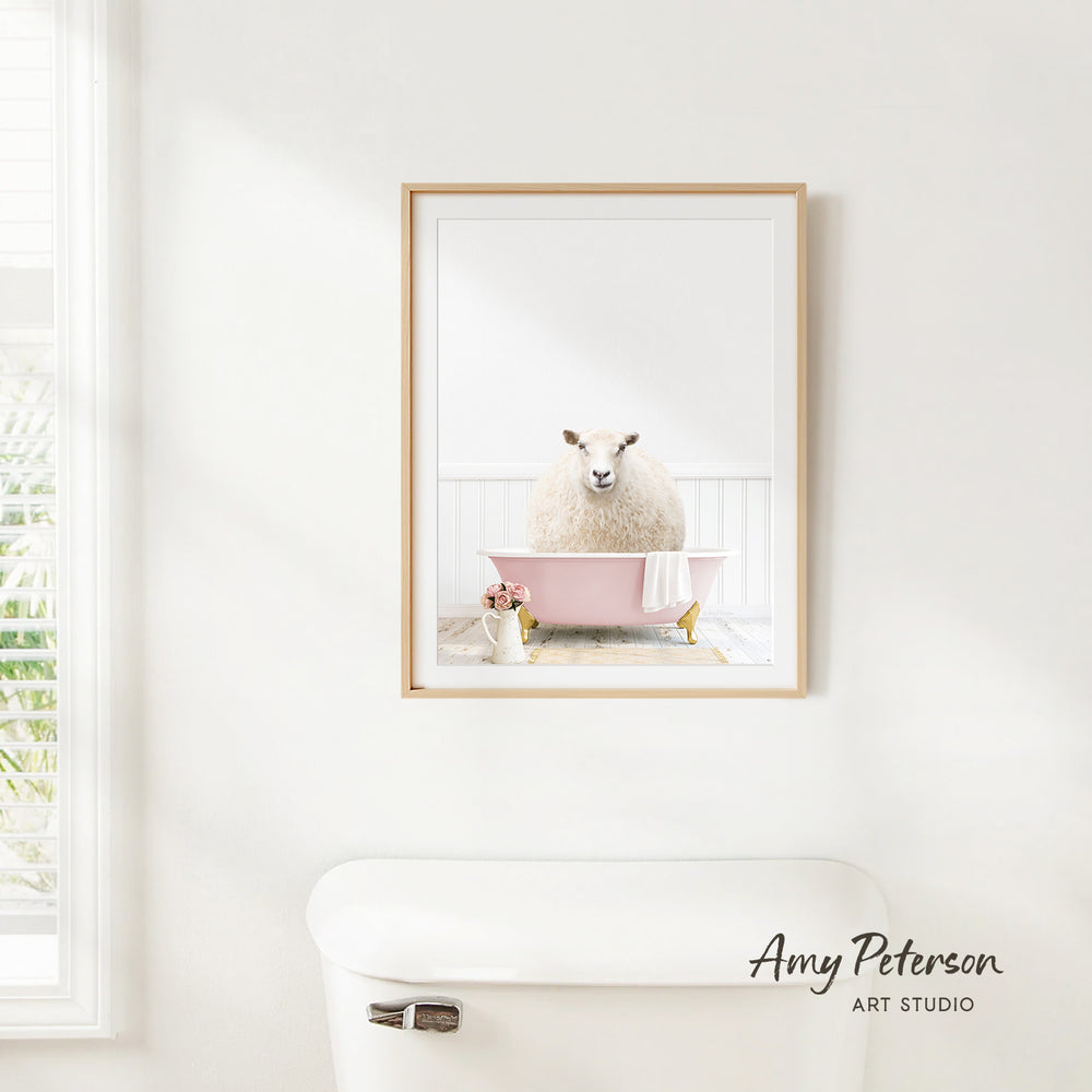 A framed image of a sheep sitting in a pink bathtub is hanging on a white wall in a bathroom.