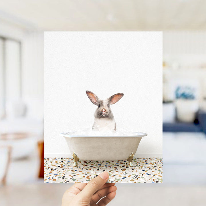 A hand holding a framed photograph of a rabbit sitting in a bathtub filled with water.