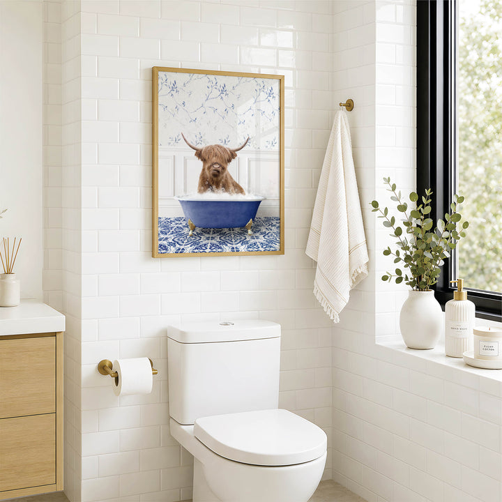 A bathroom with a white toilet, a framed picture of a Highland cow in a bathtub, and a window with greenery outside.