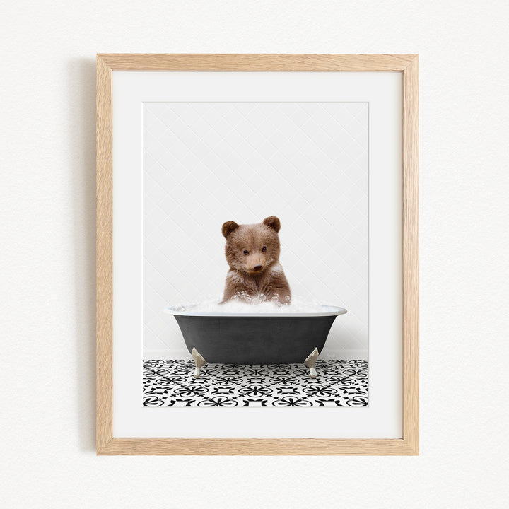 A cute brown bear cub sitting in a black bathtub filled with water, surrounded by a black and white patterned tile floor.