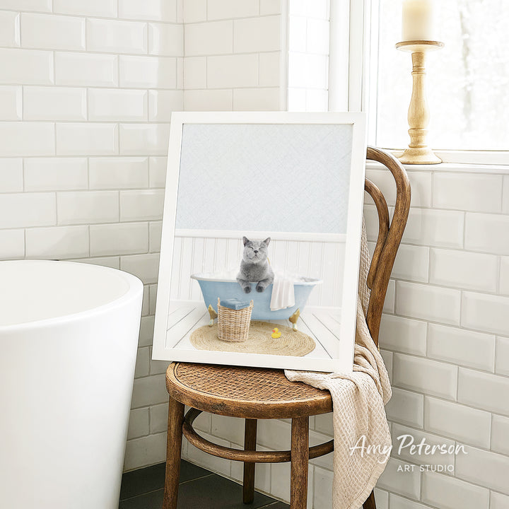 A framed image of a gray cat sitting in a blue bathtub, with a basket of bath toys nearby, is displayed on a wooden chair in a bathroom setting.