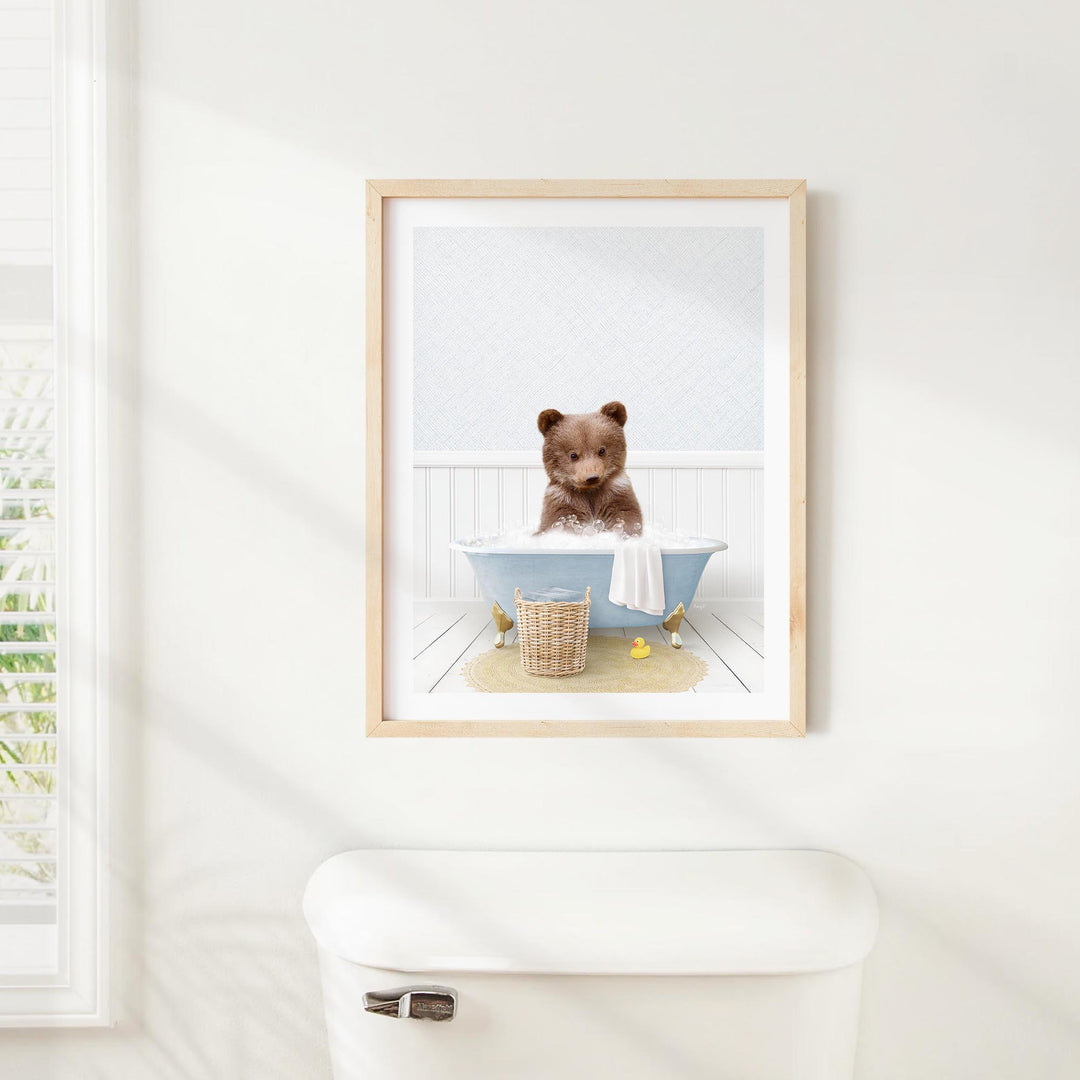 A framed image of a brown bear sitting in a blue bathtub, with a basket of bath toys nearby.