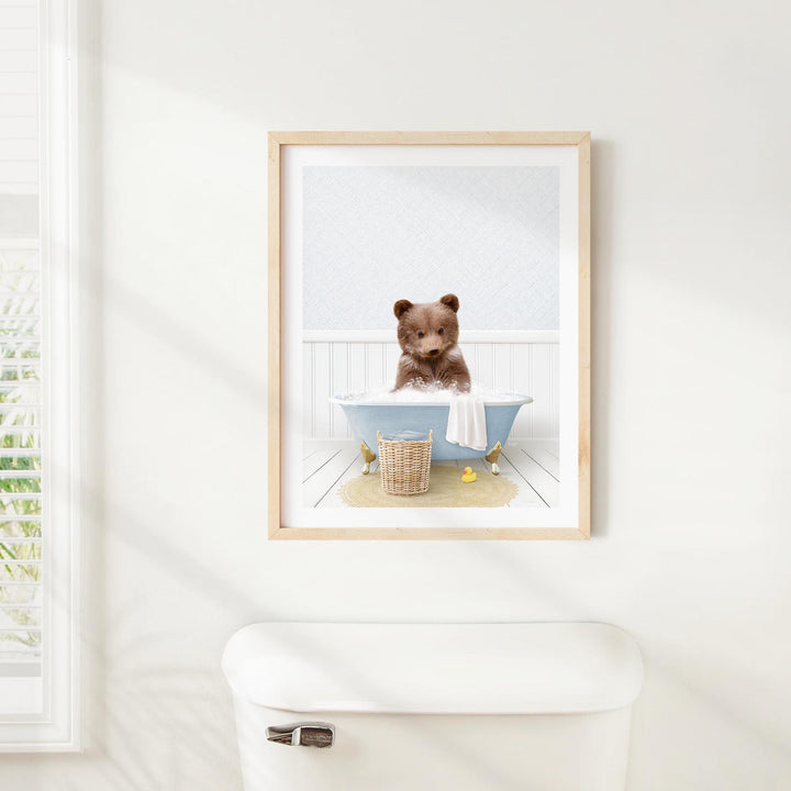 A framed image of a brown bear sitting in a blue bathtub, with a basket of bath toys nearby.