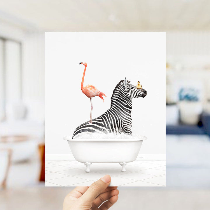 A hand holds up a framed print featuring a zebra standing in a bathtub with a pink flamingo on its back.