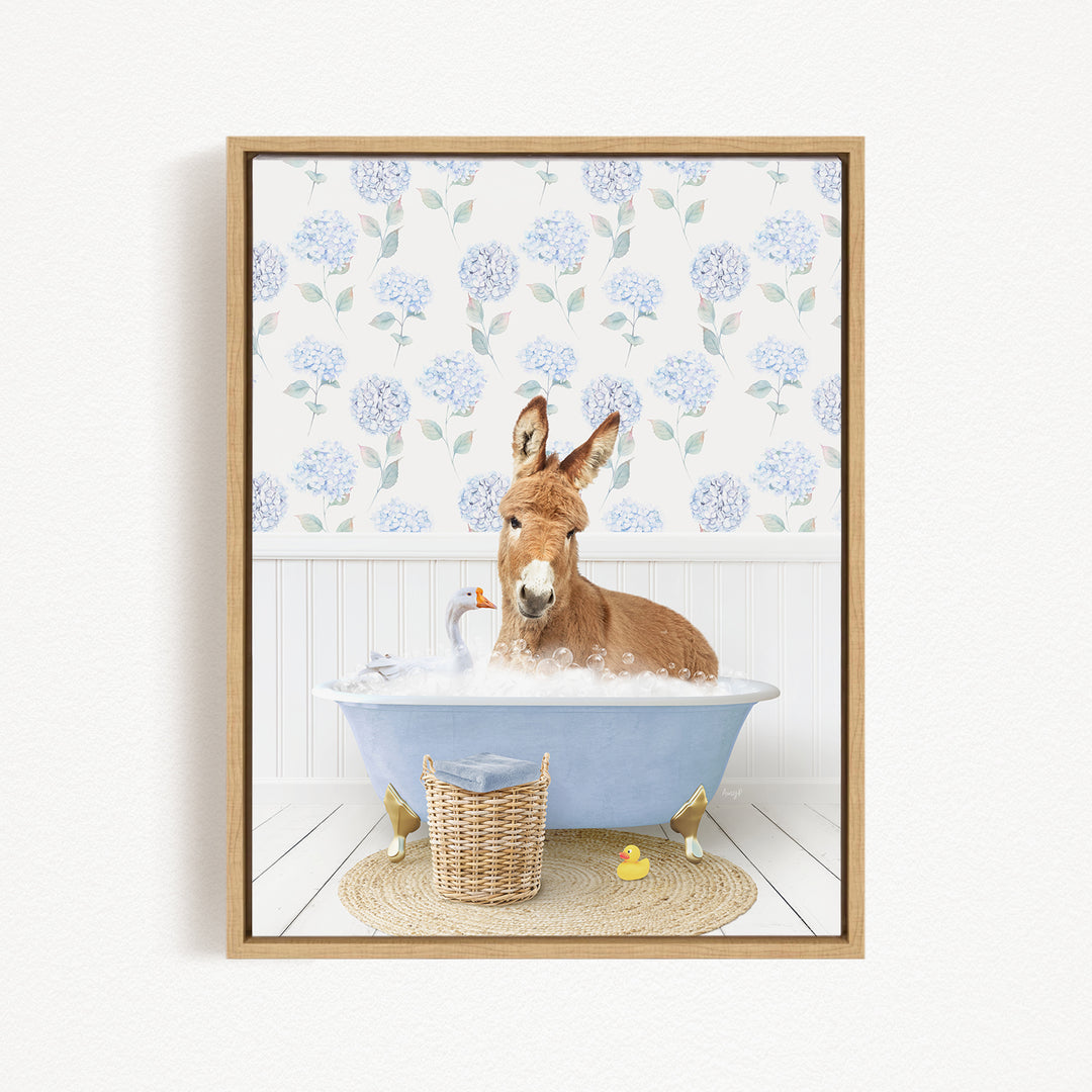 A donkey is sitting in a blue bathtub filled with bubbles, surrounded by a wicker basket and rubber duckies, with a floral patterned wall behind it.
