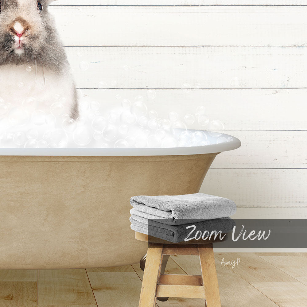 A fluffy white rabbit sitting in a bathtub filled with bubbles, with a wooden stool and towels nearby.