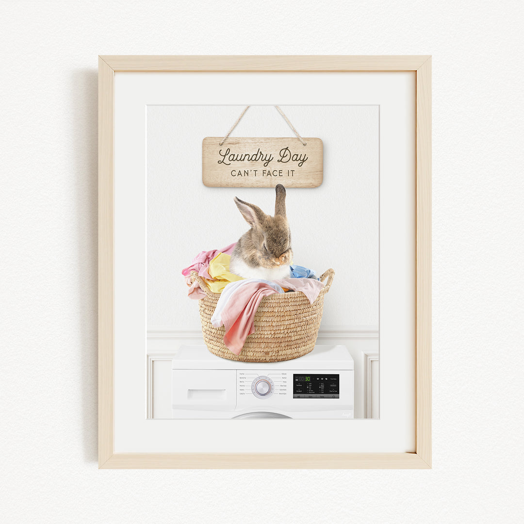 A framed image of a rabbit sitting in a basket filled with laundry, with a sign above that says "Laundry Day Can't Face It".