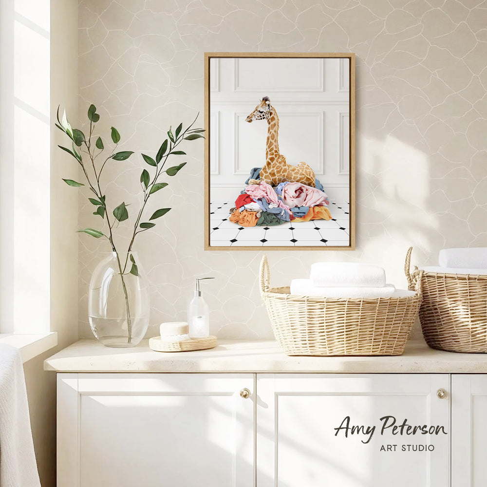 a giraffe sitting on a pile of clothes, with a framed print of the giraffe hanging on the wall above a wicker basket filled with towels.