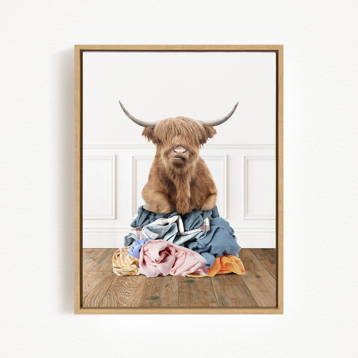 A Highland cow with long horns is sitting atop a pile of clothes on a wooden floor.