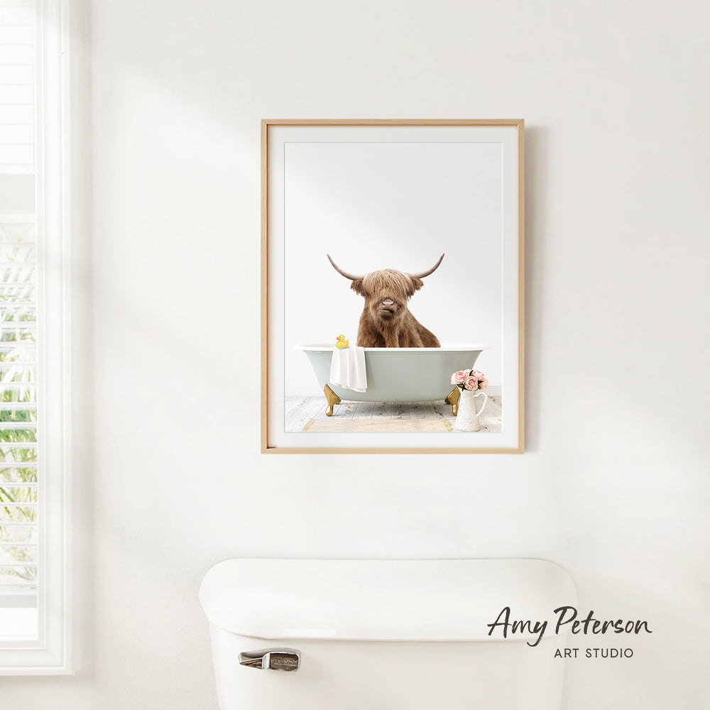 A framed image of a brown cow sitting in a bathtub, with a vase of flowers and a white toilet in the foreground.
