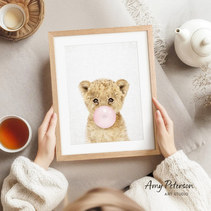 A person is holding a framed picture of a cute, fluffy animal with a pink bubblegum bubble in its mouth, surrounded by various objects such as a teapot, a cup, and a bowl.